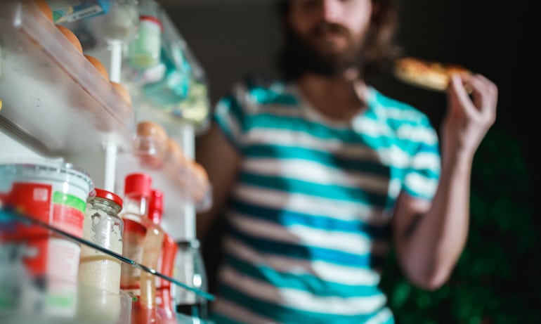 Man enjoying pizza as a midnight snack looking into the fridge because he has a late-night snacking habit