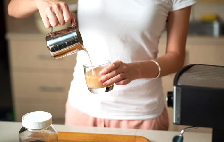 Woman making a latte at home with dairy milk, which can lead to gas and digestive discomfort on account of lactose intolerance