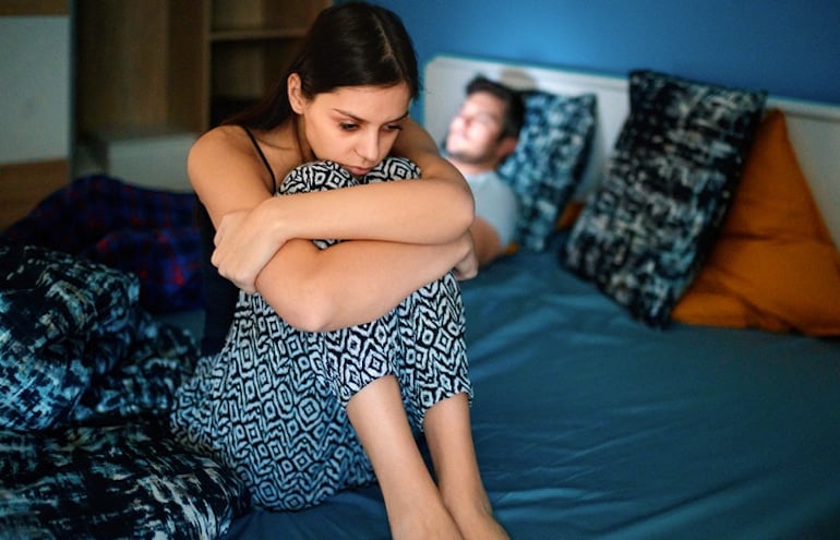 Sad woman with low libido sitting on the edge of the bed with her boyfriend unhappy about their sex life