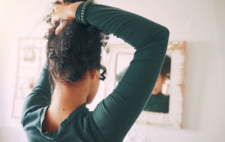 Woman looking in the mirror tying her hair up tightly, which can contribute to hair breakage