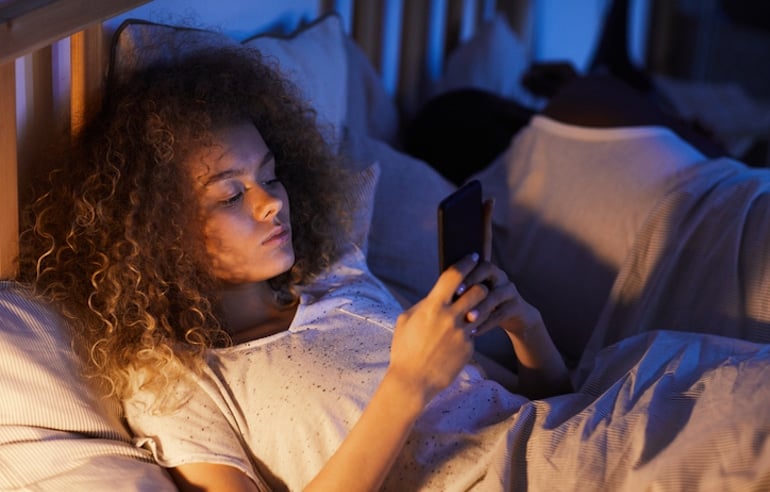 Woman using her phone in bed, which emits blue light and can lead to premature skin aging